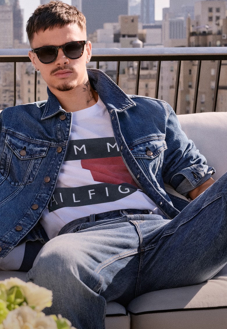 Romeo Beckham lounges on an outdoor sofa wearing a Tommy Hilfiger light denim shirt and jeans.