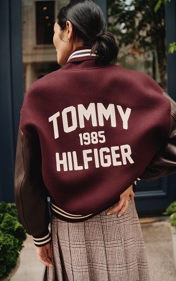 Model viewed from behind wearing a burgundy varsity jacket with "Tommy 1985 Hilfiger" across the back.