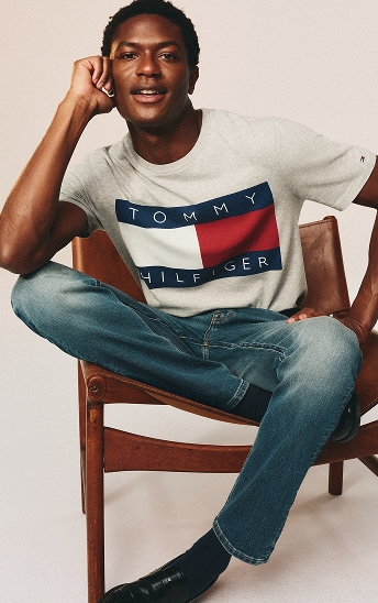 Seated model wearing a white Tommy Hilfiger logo T-shirt and light blue jeans.