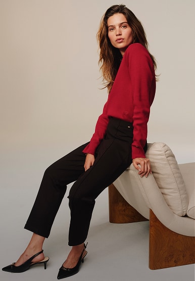 Model seated sideways on a stool wearing a Tommy Hilfiger red sweater and black tailored pants.