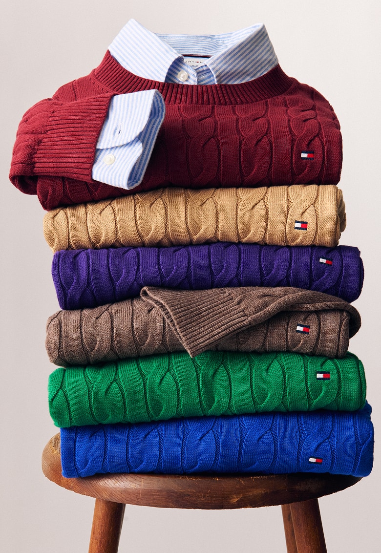 Stack of colorful Tommy Hilfiger cable-knit sweaters layered over striped button-down shirts on a wooden stool.