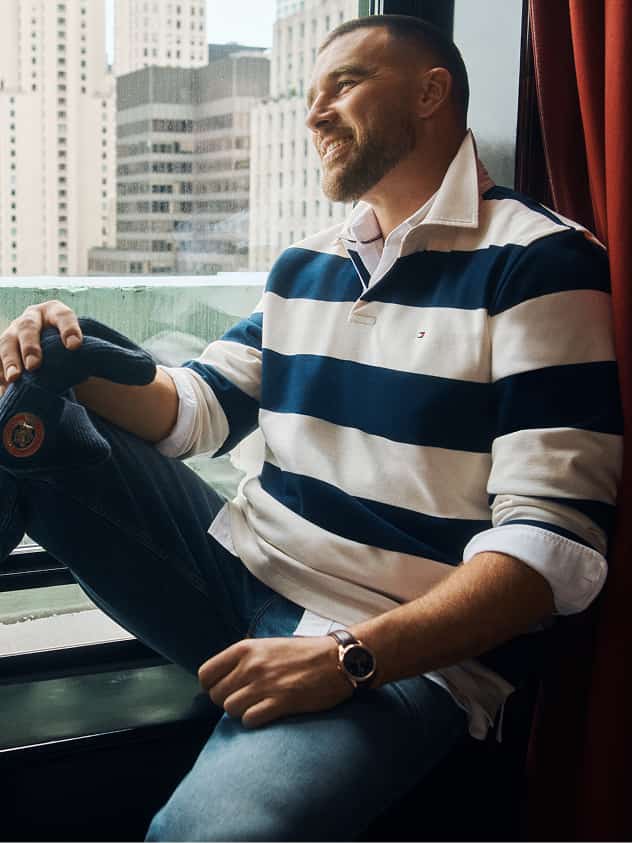 Travis Kelce wearing a Tommy Hilfiger navy and white striped rugby shirt sitting by a hotel window overlooking the city.