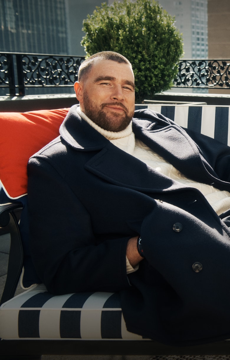 Travis Kelce in a Tommy Hilfiger navy coat and cream turtleneck relaxing on a striped outdoor sofa beside a fluffy white dog on a New York rooftop terrace.