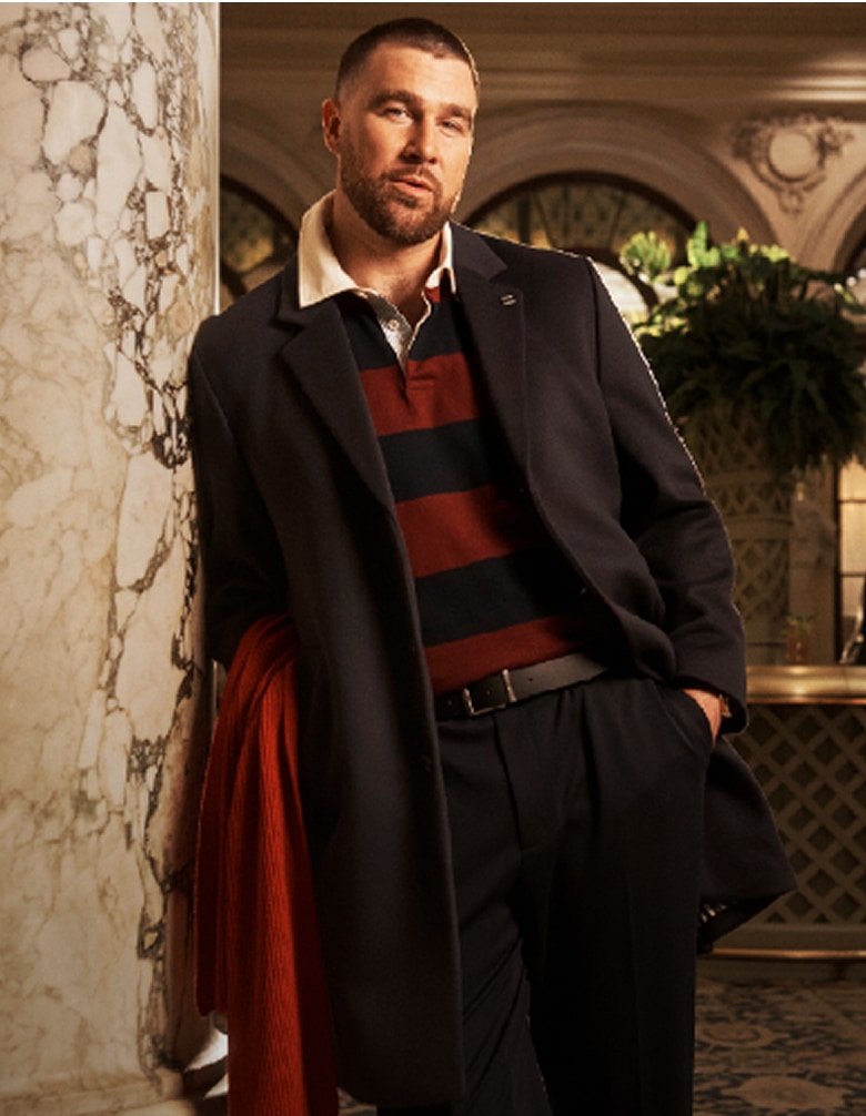 Travis Kelce wearing a Tommy Hilfiger navy overcoat over a striped rugby shirt, standing in an elegant hotel lobby.