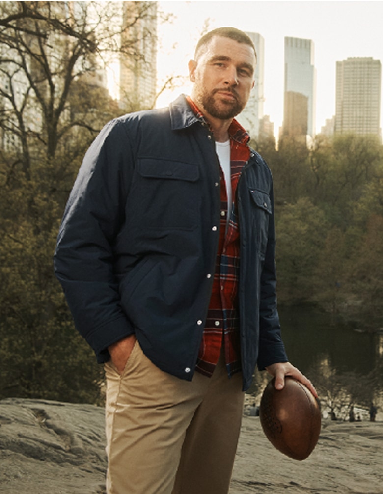 Travis Kelce wearing a Tommy Hilfiger navy overshirt layered over a plaid shirt and khaki pants, standing in Central Park with the New York skyline behind him.