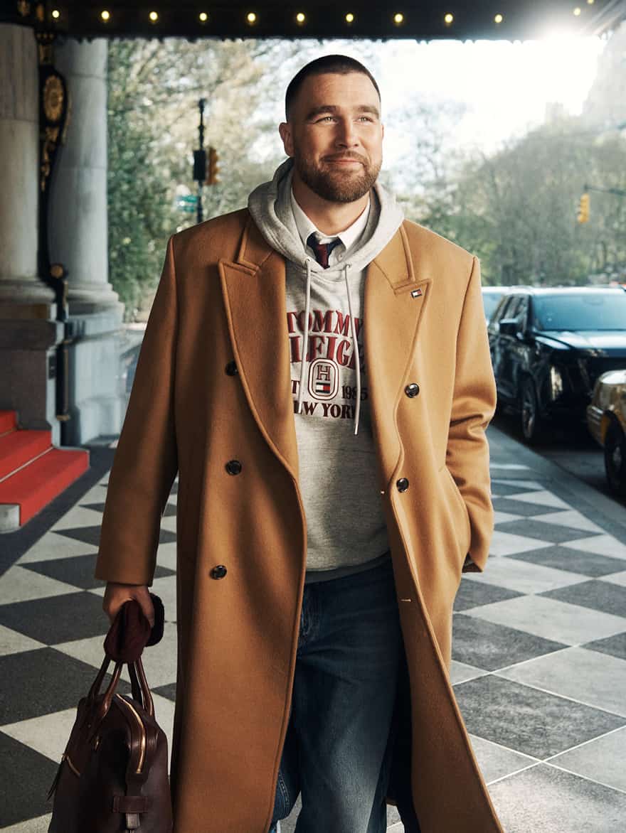 Travis Kelce wearing a Tommy Hilfiger camel double-breasted coat over a gray hoodie and jeans, standing at a building entrance.