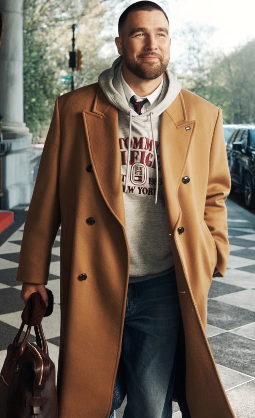 Travis Kelce wearing a Tommy Hilfiger camel double-breasted coat over a gray hoodie and jeans, standing at a building entrance.