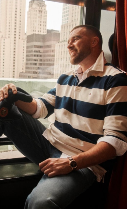Travis Kelce wearing a Tommy Hilfiger navy and white striped rugby shirt sitting by a hotel window overlooking the city.